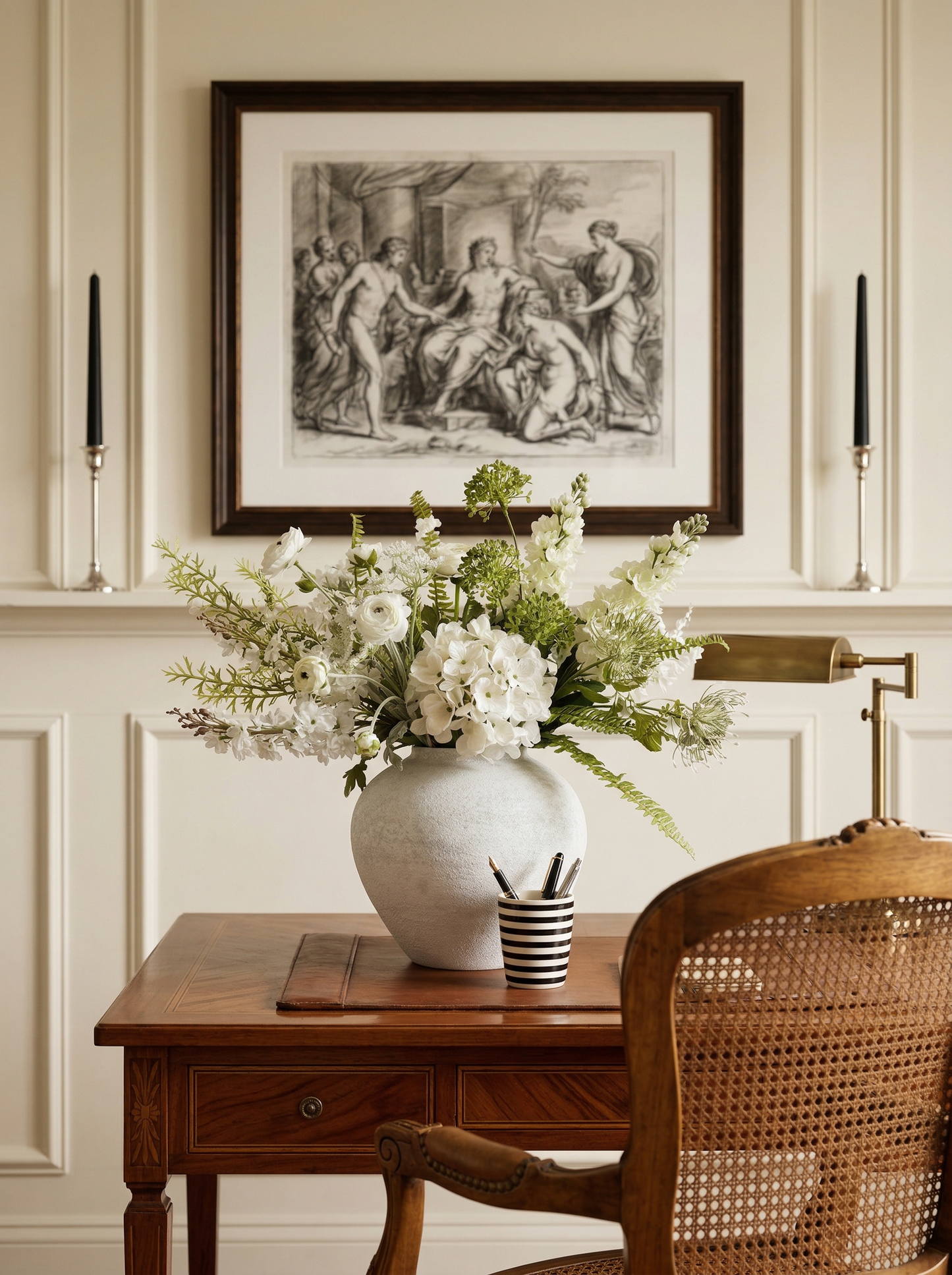 Decorative interior with a vase of flowers on a wooden table, framed artwork above, and a wicker chair.