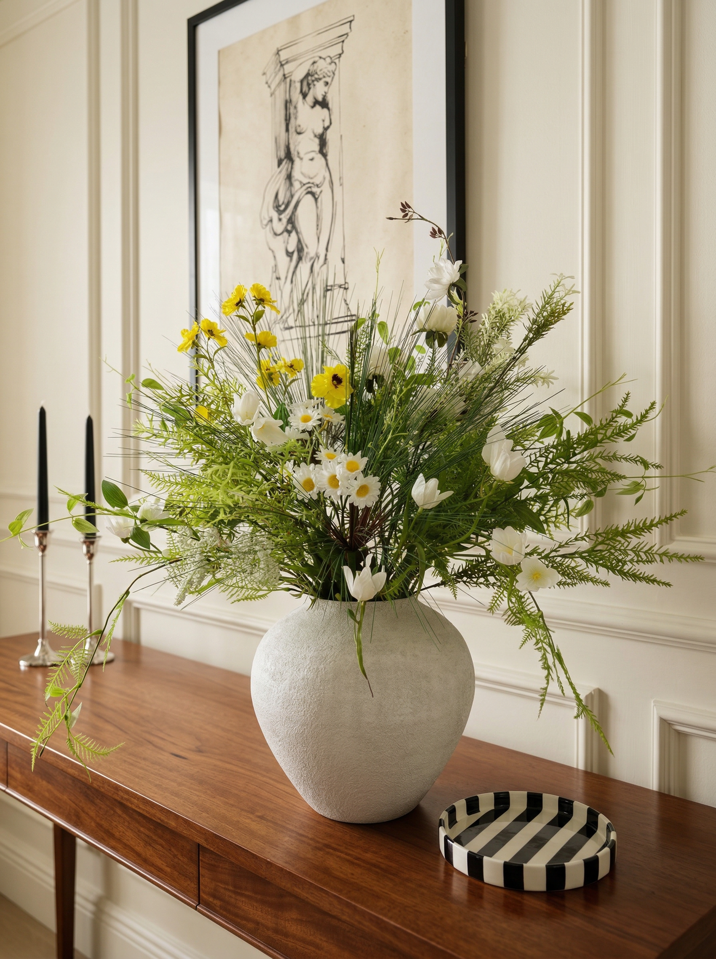 White vase with flowers on a wooden table in a room with a framed picture on the wall.