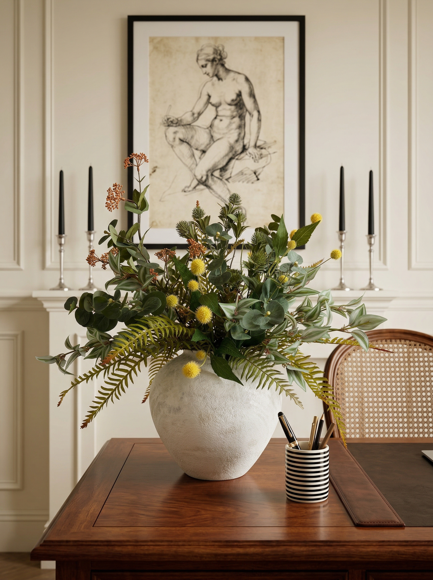Decorative floral arrangement in a white vase on a wooden table with candles and a framed artwork in the background.