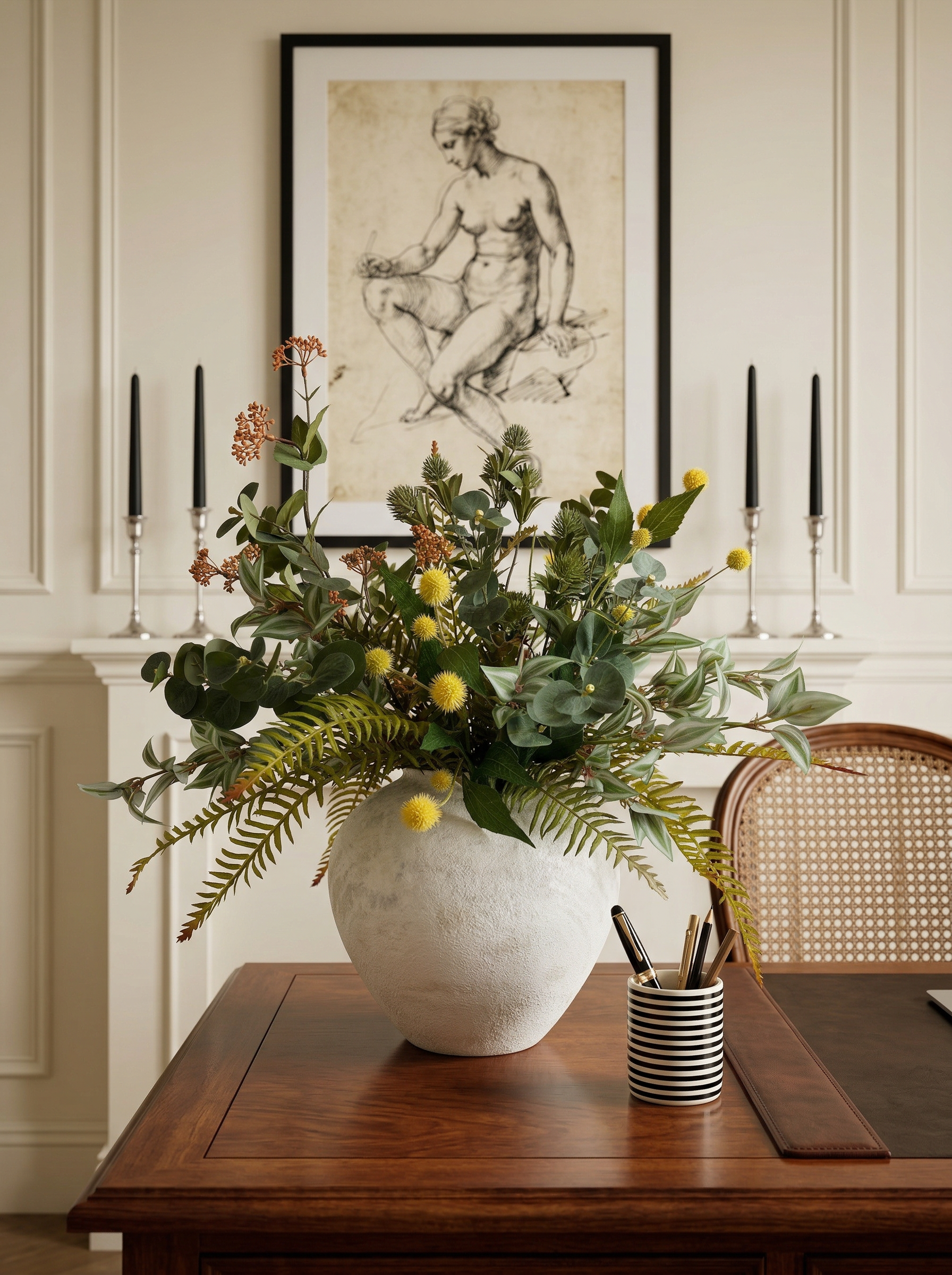 Decorative floral arrangement in a white vase on a wooden table with candles and a framed artwork in the background.