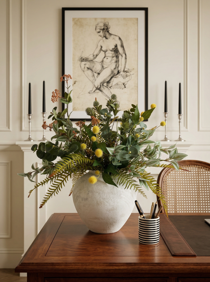 Decorative floral arrangement in a white vase on a wooden table with candles and a framed artwork in the background.