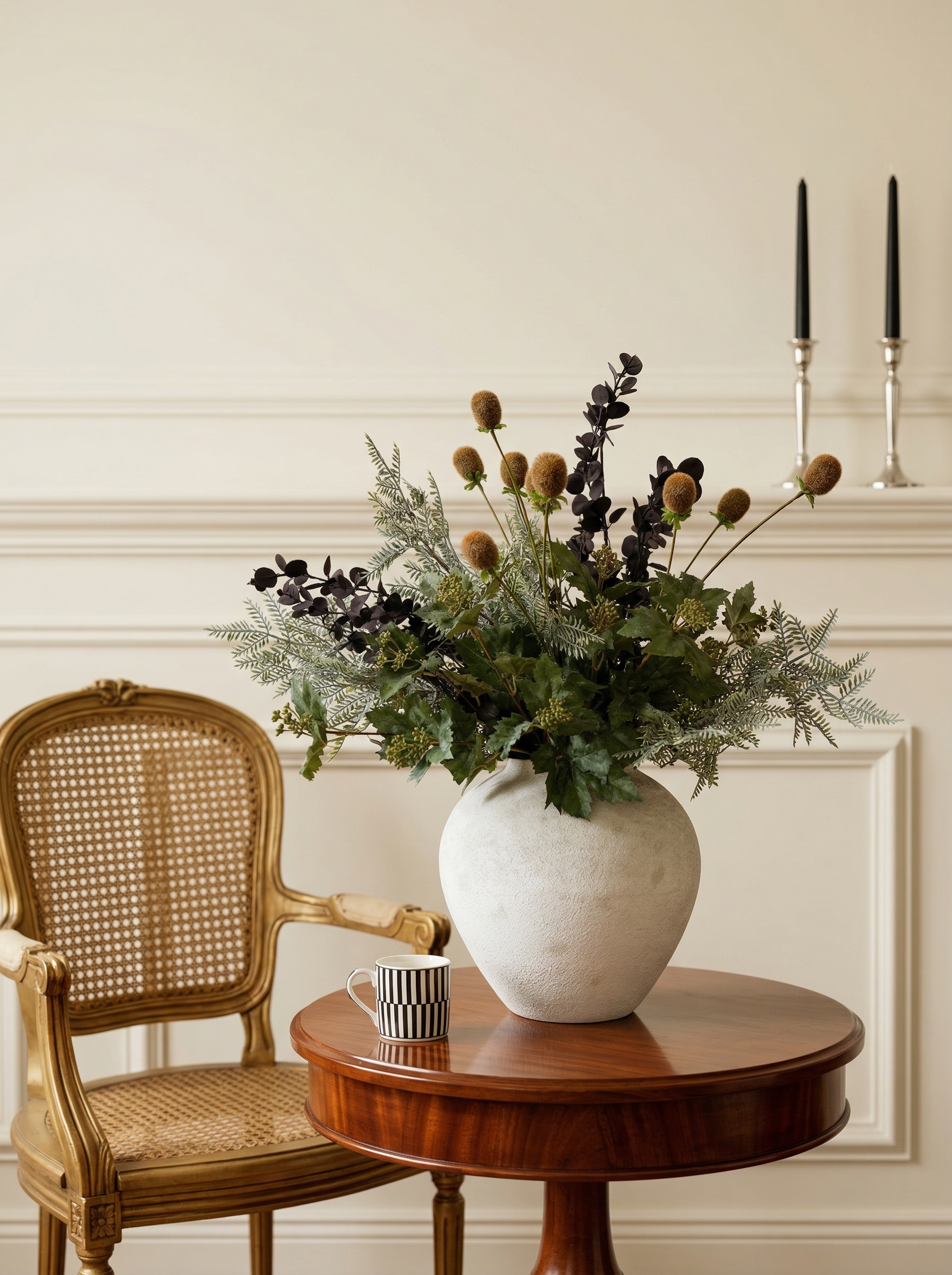 Decorative arrangement of flowers in a white vase on a wooden table with a wicker chair and candles in the background.