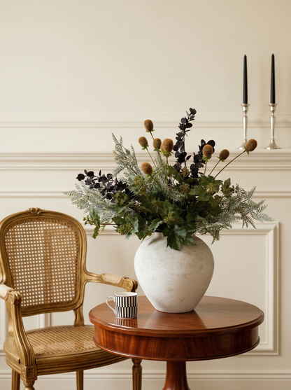 Decorative arrangement of flowers in a white vase on a wooden table with a wicker chair and candles in the background.