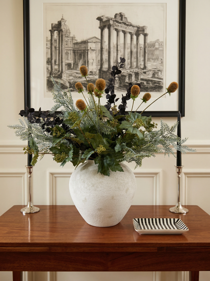 Decorative floral arrangement in a white vase on a wooden table with a framed black and white print in the background.
