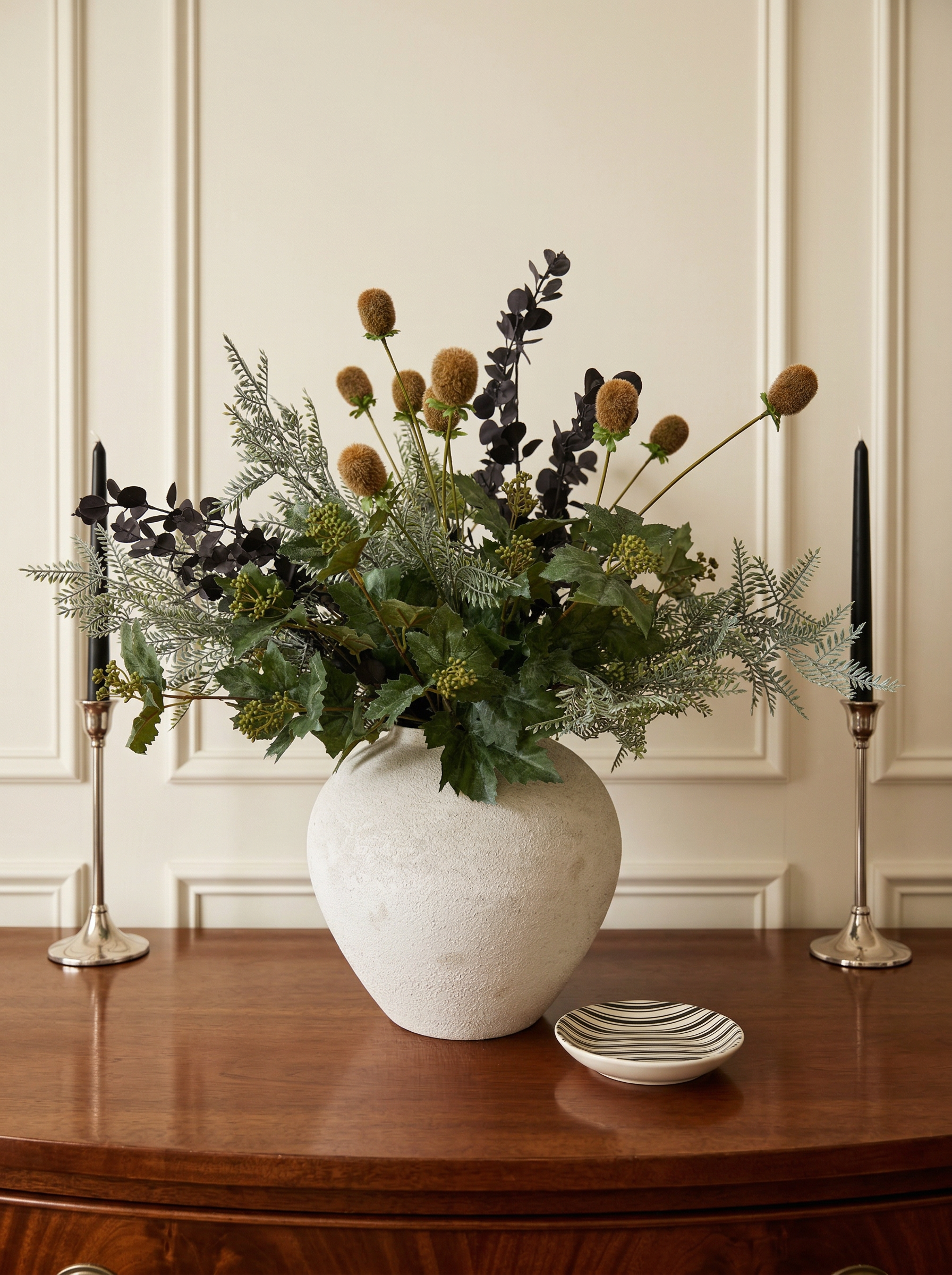 Decorative floral arrangement in a white vase on a wooden table with candles and a plate.
