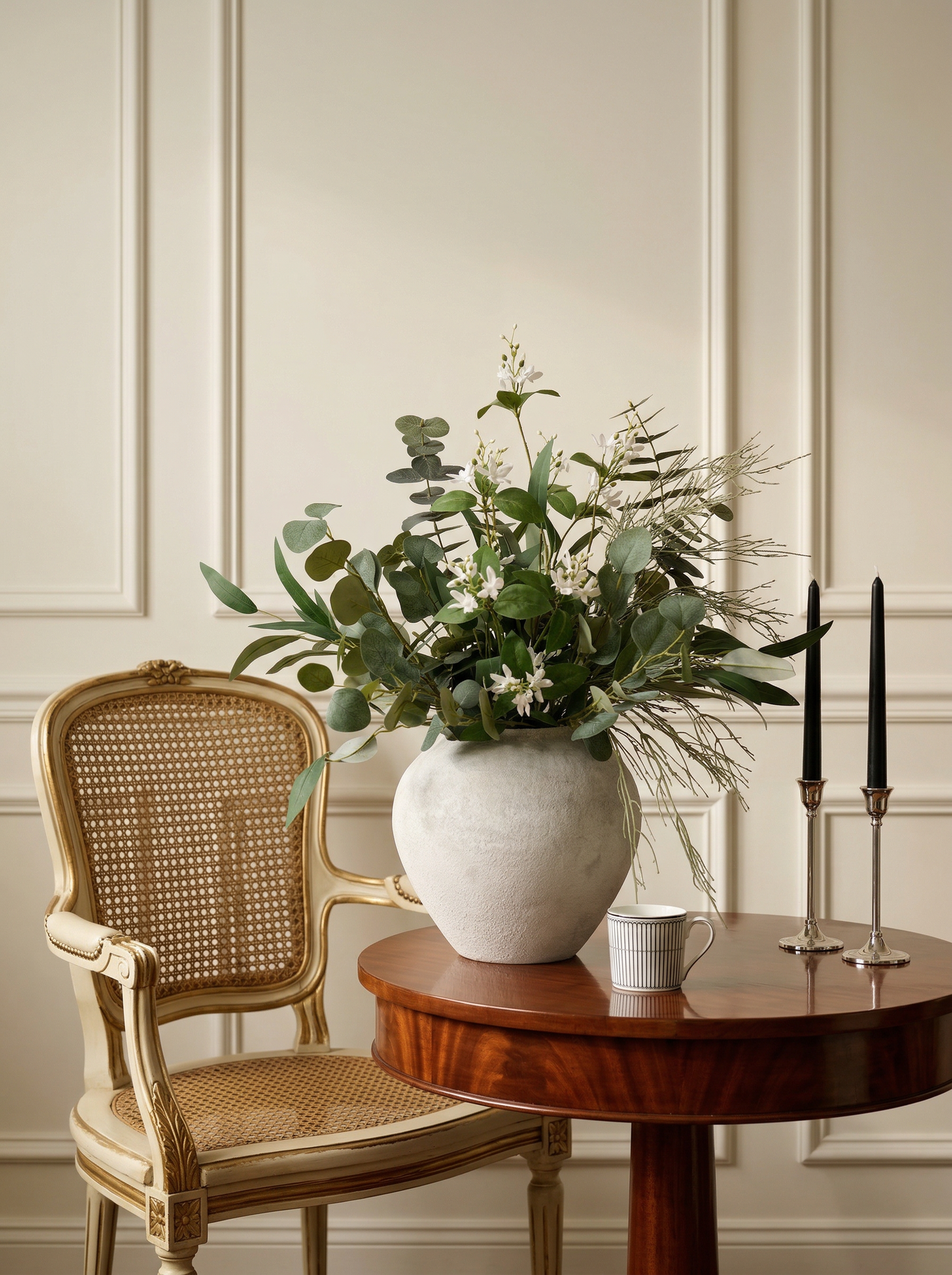 Decorative setting with a vase of greenery, candles, and a chair against a paneled wall.