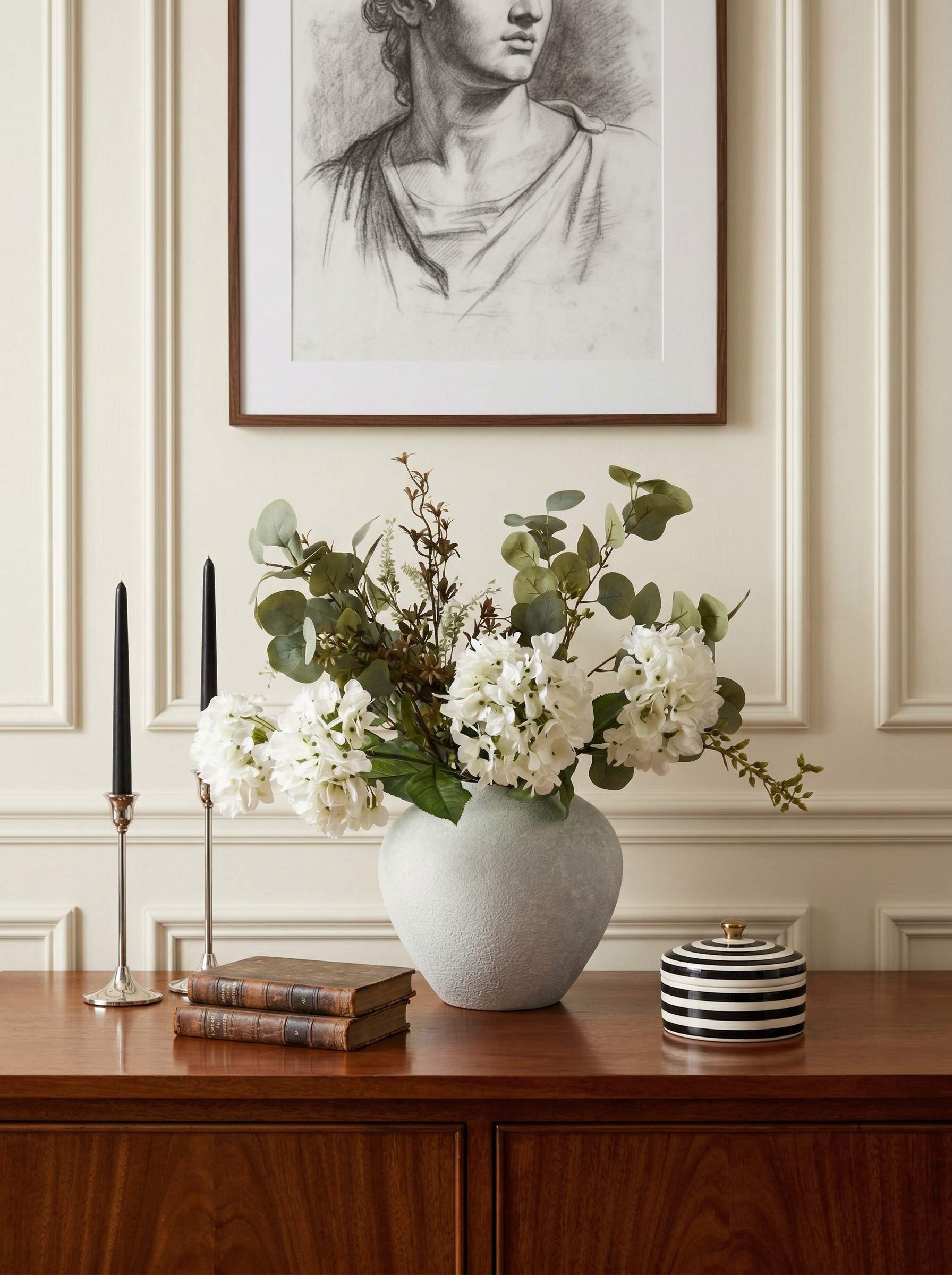Decorative setup with a vase of flowers, books, and candles on a wooden surface, with a framed portrait in the background.