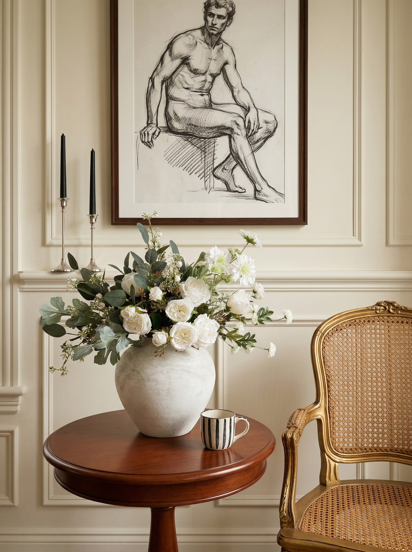 Floral arrangement in a vase on a wooden table with a wicker chair and framed artwork in the background.