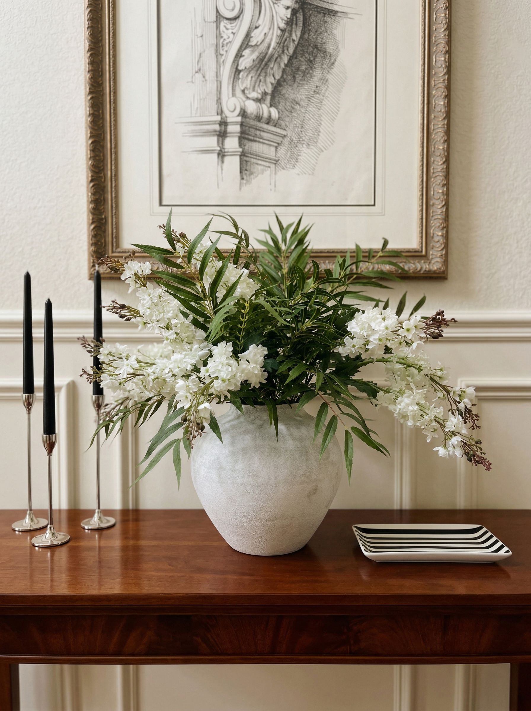 Floral arrangement in a vase on a wooden table with candles and a framed picture in the background.