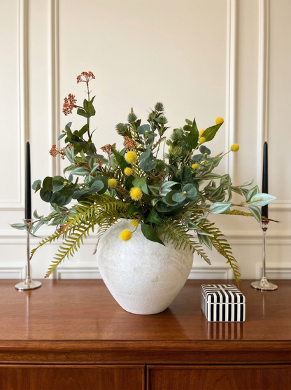 Floral arrangement in a white vase on a wooden surface with candles and a box in the background.