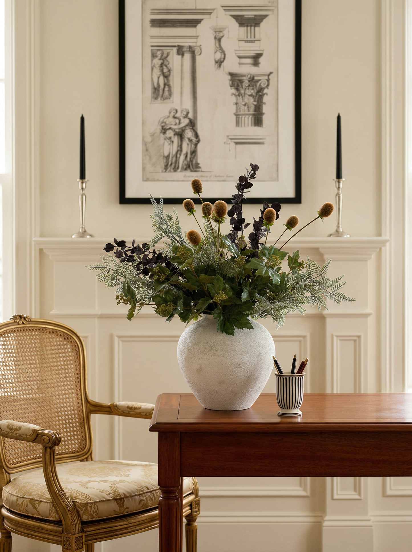 Floral arrangement in a white vase on a wooden table with a chair and framed artwork in the background.