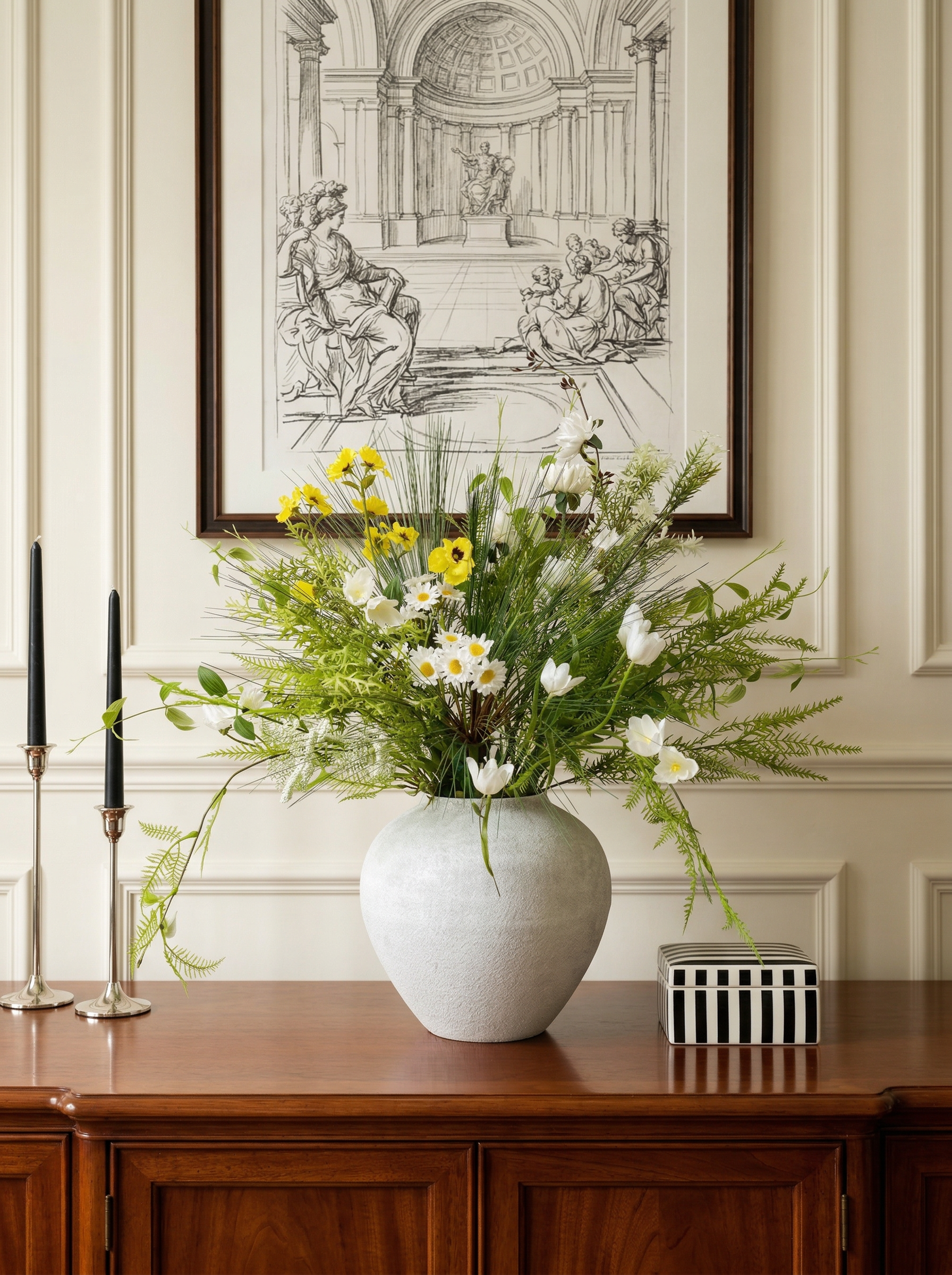 Floral arrangement in a white vase on a wooden table with a framed black and white artwork in the background.