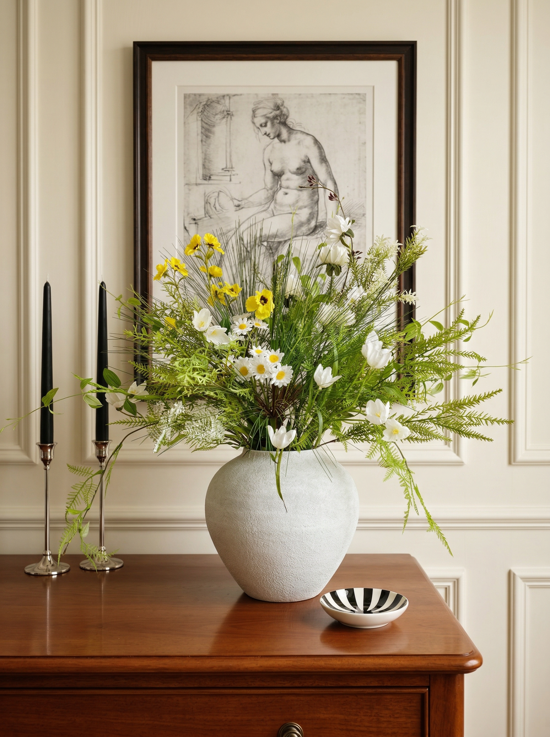 Floral arrangement in a white vase on a wooden table with candles and a bowl in a room with a framed artwork on the wall.