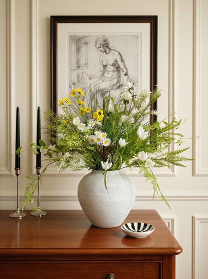 Floral arrangement in a white vase on a wooden table with candles and a bowl in a room with a framed artwork on the wall.