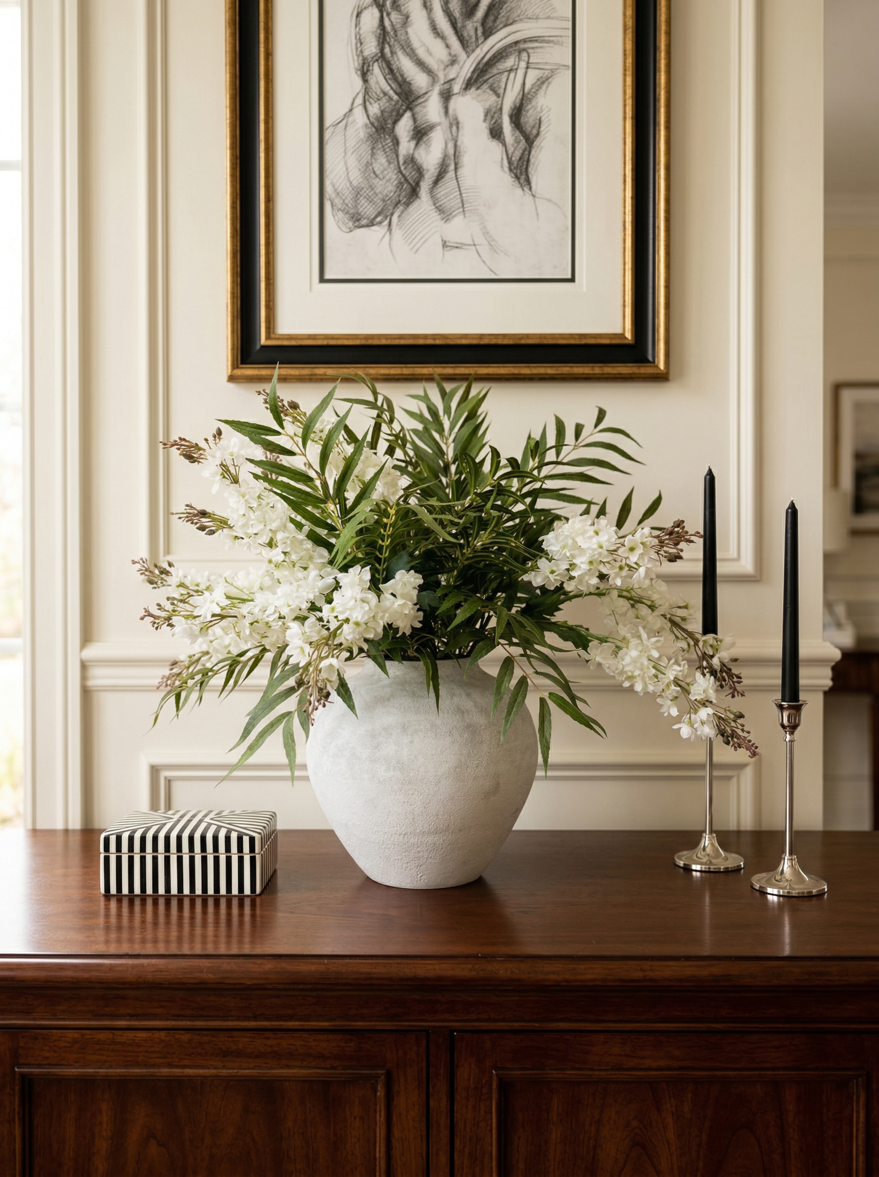 Floral arrangement in a white vase on a wooden table with framed artwork in the background.