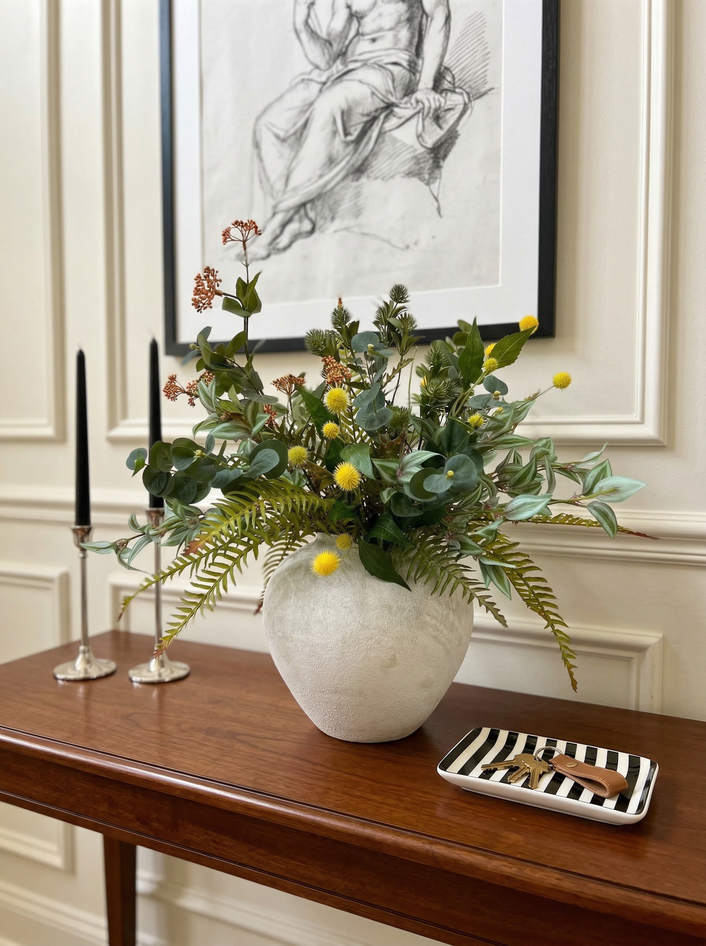Vase with greenery on a wooden table with candles and a decorative plate.