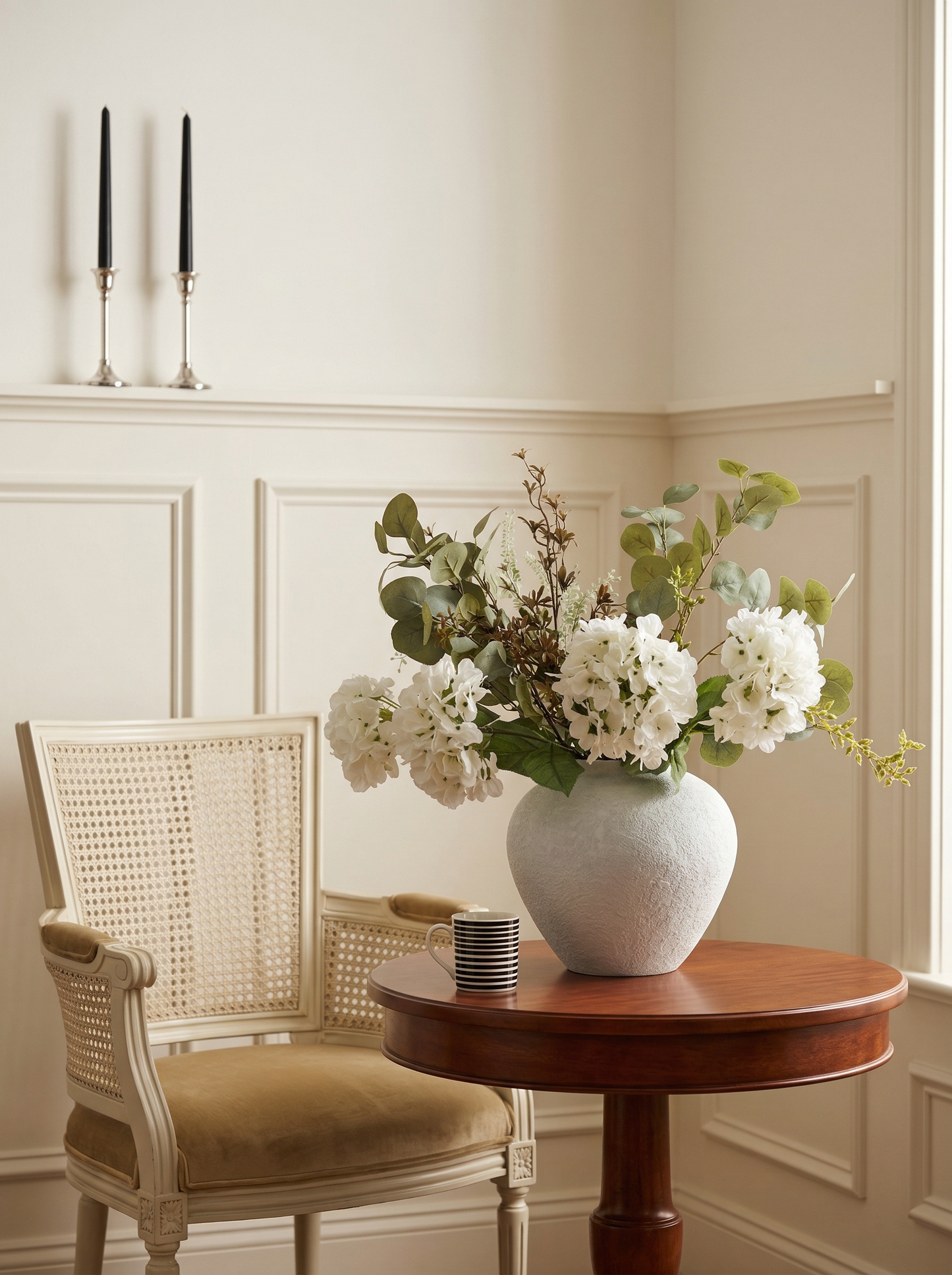 Vase with white flowers on a wooden table in a room with a chair and candles.